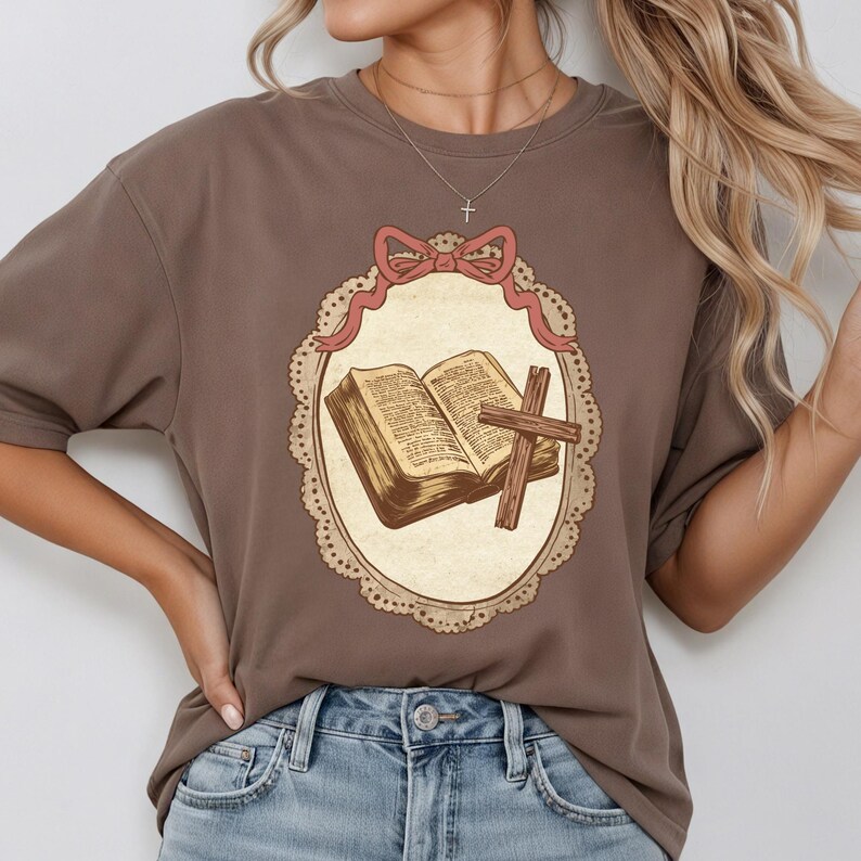 May include: A brown t-shirt featuring a framed illustration of an open book and a cross. The illustration is in a cream-colored oval with a pink bow and a decorative border. The t-shirt is a solid brown color.
