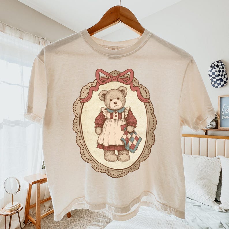 Op de afbeelding: Cr&egrave;mekleurig T-shirt met een vintage illustratie van een teddybeer in een jurk, met een quilt. De beer is omlijst in een ovale rand met een roze strik. Het T-shirt hangt aan een houten hanger.