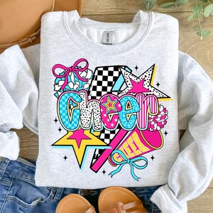 Op de afbeelding: Een lichtgrijs sweatshirt met een kleurrijke "Cheer"-afbeelding. Het ontwerp bevat sterren, een megafoon en een strik, met het woord "Cheer" in een mix van lettertypen en patronen, waaronder geblokte en luipaardprint.