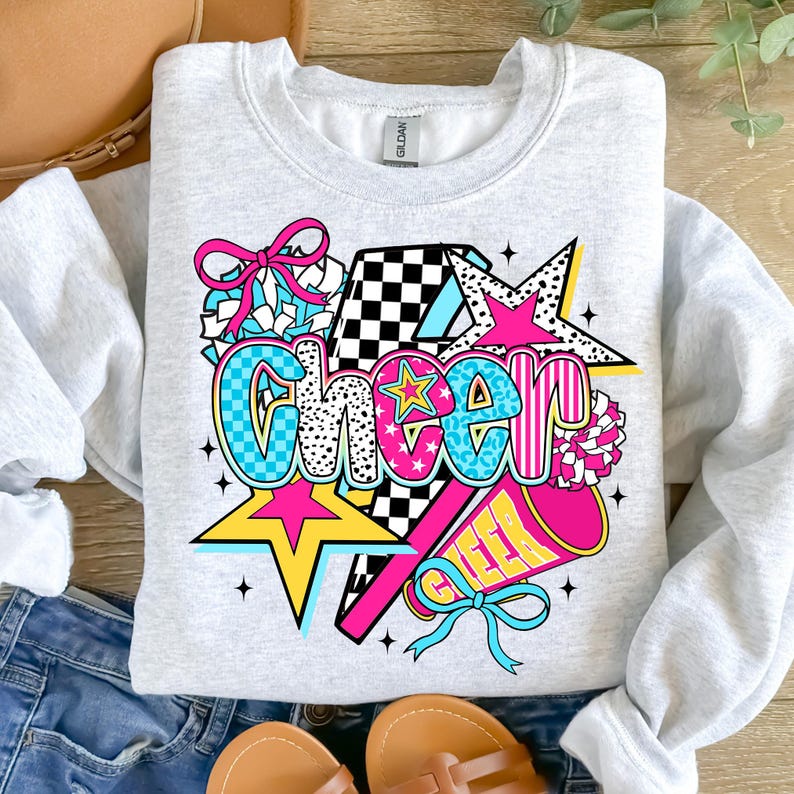Op de afbeelding: Een lichtgrijs sweatshirt met een kleurrijke "Cheer"-afbeelding. Het ontwerp bevat sterren, een megafoon en een strik, met het woord "Cheer" in een mix van lettertypen en patronen, waaronder geblokte en luipaardprint.