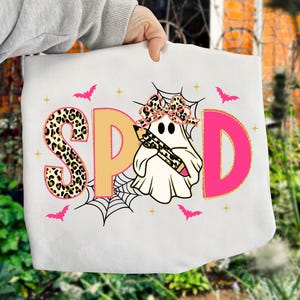 Pu&ograve; includere: Borsa tote bianca con la scritta "SPD" in un design giocoso. La lettera "S" ha un motivo leopardato, mentre la "D" &egrave; rosa. Un fantasma con un fiocco leopardato e una matita &egrave; al centro, con pipistrelli e ragnatele.