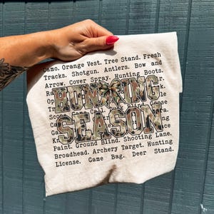 May include: A beige fabric item with the words "HUNTING SEASON" in large, camouflage-patterned letters. The text is surrounded by a list of hunting-related terms. A camouflage bow adds a decorative touch.