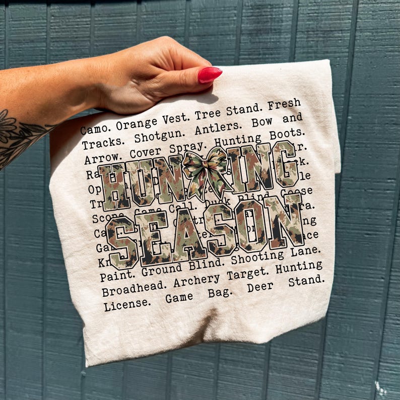 May include: A beige fabric item with the words "HUNTING SEASON" in large, camouflage-patterned letters. The text is surrounded by a list of hunting-related terms. A camouflage bow adds a decorative touch.