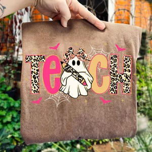 May include: Brown t-shirt with the word "TEACH" in pink and leopard print. A ghost wearing a leopard print bow and holding a pencil is in the center. Pink bats and spiderwebs complete the Halloween-themed design.