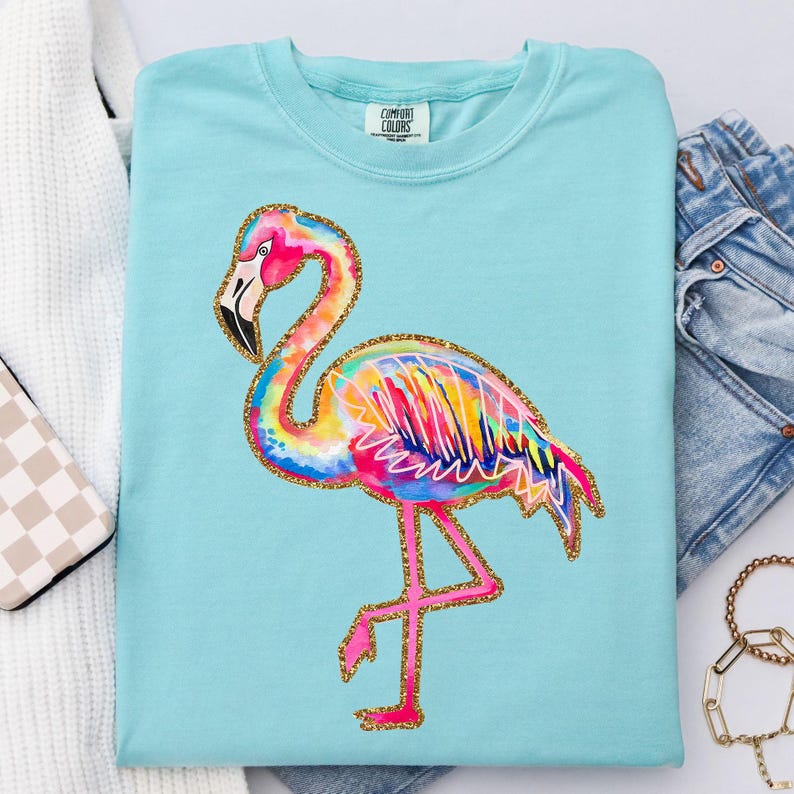 May include: A light blue Comfort Colors t-shirt featuring a colorful flamingo design. The flamingo has a pink head and legs, a black beak, and a body with rainbow hues. Gold glitter outlines the bird. The shirt is folded.