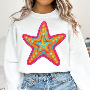 May include: White sweatshirt featuring a vibrant, colorful starfish design. The starfish is outlined in hot pink, with orange, yellow, and blue interior details. The design includes small yellow circles along the starfish's edges. The sweatshirt is paired with blue denim jeans.
