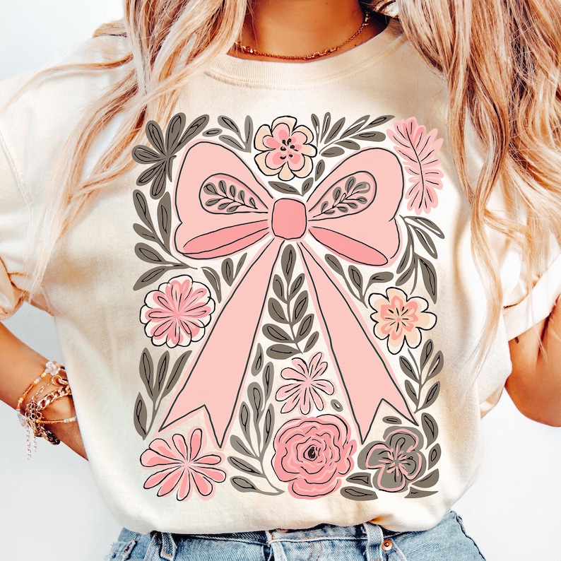 K&ouml;nnte beinhalten: Cremefarbenes Sweatshirt mit einer gro&szlig;en rosa Schleife, umgeben von floralen und Blattillustrationen in Rosa, Grau und Wei&szlig;. Das Design ist auf der Vorderseite zentriert und schafft eine feminine und k&uuml;nstlerische &Auml;sthetik.
