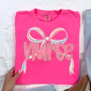 Pode incluir: Uma t-shirt rosa brilhante com a palavra "Nurse" em uma fonte l&uacute;dica, acentuada por um design de la&ccedil;o colorido. O la&ccedil;o apresenta tons pastel arco-&iacute;ris, adicionando um toque alegre ao design da t-shirt. A t-shirt est&aacute; cuidadosamente dobrada.