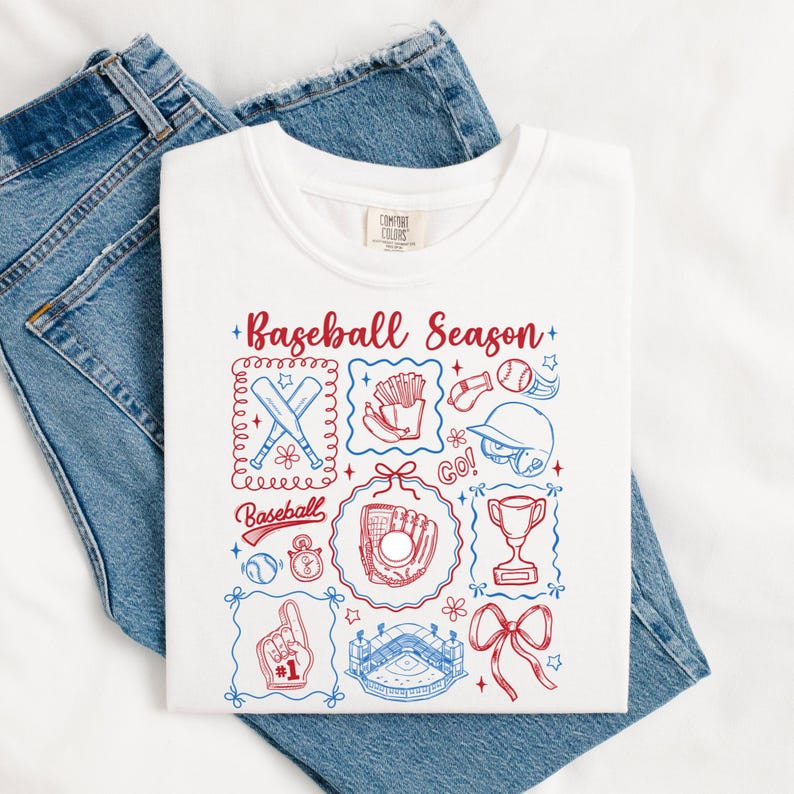 May include: White t-shirt with a baseball-themed graphic. The design includes baseball bats, a glove, a stadium, and the text "Baseball Season" in red and blue. The shirt is laid on top of blue jeans.