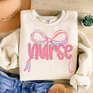 Pode incluir: Camisola creme com um grande la&ccedil;o colorido e a palavra "Nurse" em rosa. O la&ccedil;o apresenta listras de arco-&iacute;ris pastel. A camisola &eacute; um estilo casual e confort&aacute;vel.