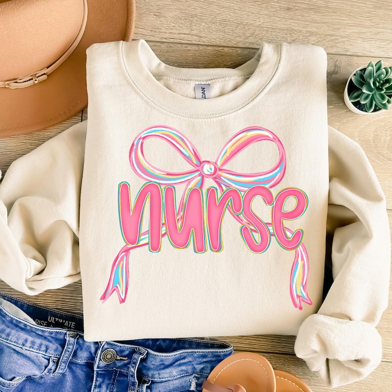 Pode incluir: Camisola creme com um grande la&ccedil;o colorido e a palavra "Nurse" em rosa. O la&ccedil;o apresenta listras de arco-&iacute;ris pastel. A camisola &eacute; um estilo casual e confort&aacute;vel.