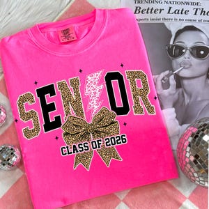 Pu&ograve; includere: Felpa rosa acceso con la scritta "SENIOR" in stampa leopardata e lettere nere, un fiocco con stampa leopardata e la scritta "CLASS OF 2026". La maglietta presenta anche un fulmine rosa e bianco.
