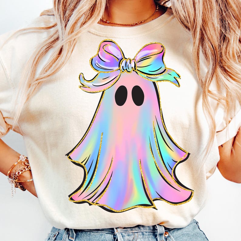 May include: A cream-colored t-shirt featuring a colorful ghost design. The ghost has a rainbow-colored body, black eyes, and a large bow on its head. The design is outlined in gold glitter.