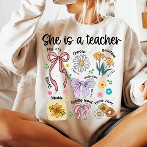 Op de afbeelding: Cr&egrave;mekleurige sweatshirt met de tekst "She is a teacher" en bloemmotieven. Het ontwerp bevat een roze strik, madeliefjes en andere bloemen, samen met inspirerende woorden als "Chosen" en "Victorious".