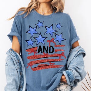 Pode incluir: Uma camiseta azul com um design de glitter vermelho, branco e azul de estrelas e uma bandeira com a palavra "AND" em preto.