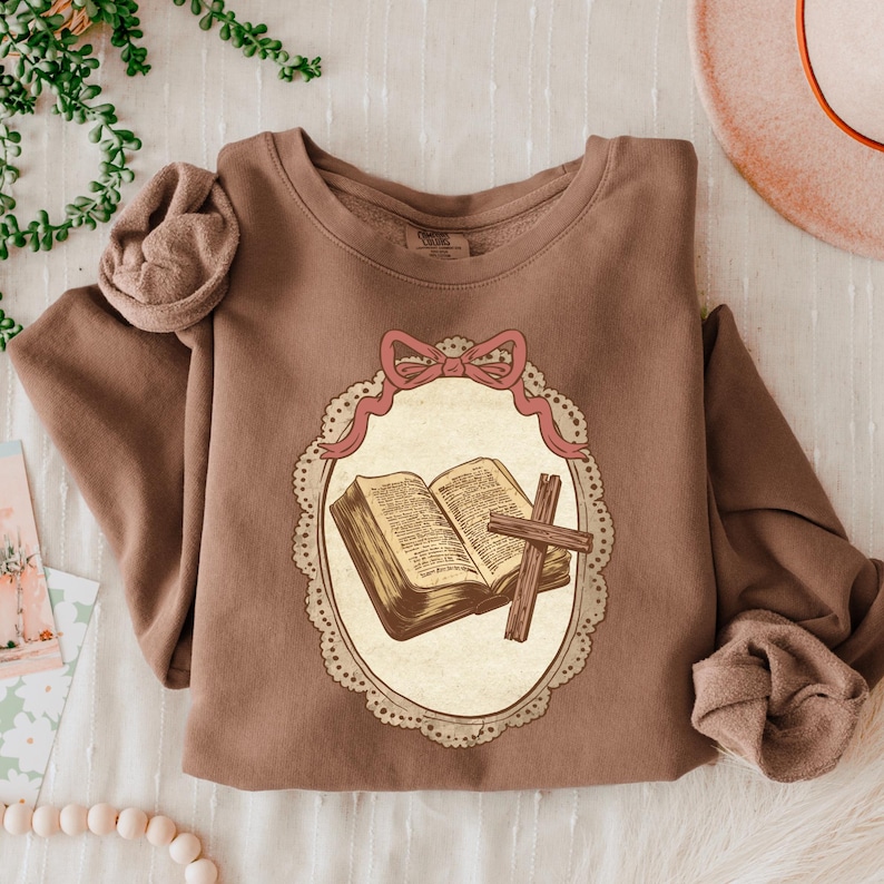 May include: A brown sweatshirt featuring a graphic of an open book and a cross within a decorative oval frame. The frame is adorned with a pink bow and ribbon. The sweatshirt has long sleeves.