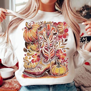 Op de afbeelding: Witte sweatshirt met een aquarelontwerp van een cowboylaars, pompoenen en herfstbladeren in bruin, oranje en rood. De laars staat centraal, omringd door bloemen en pompoenaccenten, wat een seizoensgebonden thema cre&euml;ert.