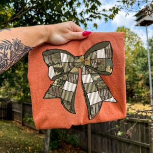 May include: An orange-colored t-shirt featuring a large bow design. The bow is a patchwork of camouflage and plaid patterns in shades of green, brown, and white. The shirt is held up against a background of greenery and a blue sky.