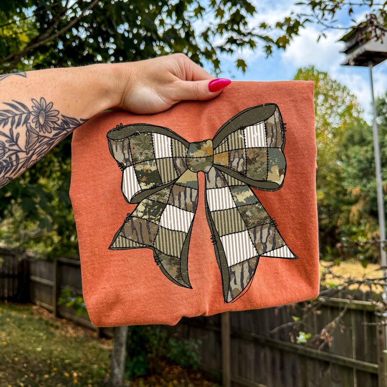 May include: An orange-colored t-shirt featuring a large bow design. The bow is a patchwork of camouflage and plaid patterns in shades of green, brown, and white. The shirt is held up against a background of greenery and a blue sky.