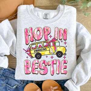 May include: A gray sweatshirt with a pink and gold graphic design. The graphic features a yellow school bus with the words "Struggle Bus" on the side. The text "Hop in bestie" is written in pink and gold letters above the bus.