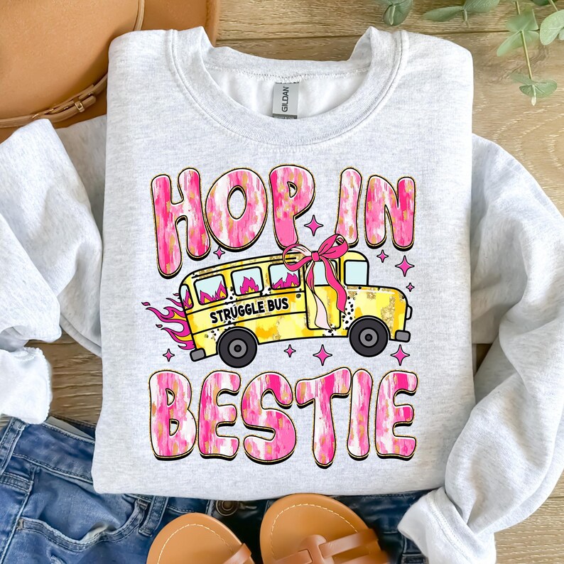 May include: A gray sweatshirt with a pink and gold graphic design. The graphic features a yellow school bus with the words "Struggle Bus" on the side. The text "Hop in bestie" is written in pink and gold letters above the bus.