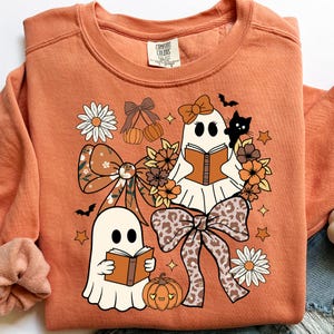 Op de afbeelding: Oranje Comfort Colors sweatshirt met een grillig Halloween-ontwerp. Twee spookfiguren lezen boeken, omringd door strikken, bloemen, pompoenen, sterren en een zwarte kat. Het ontwerp heeft een mix van herfstkleuren.