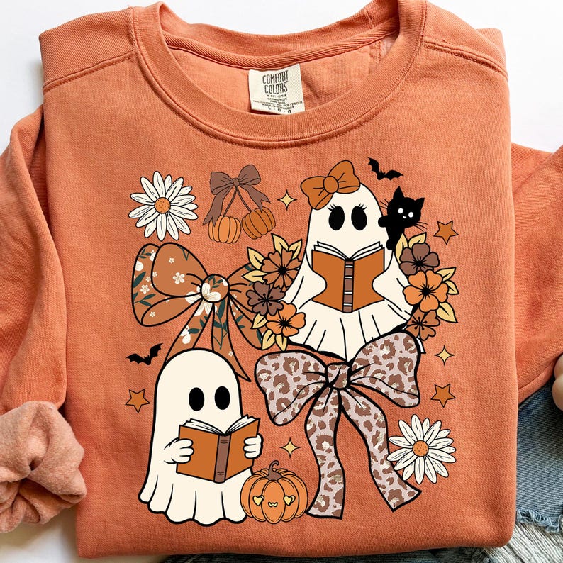 Op de afbeelding: Oranje Comfort Colors sweatshirt met een grillig Halloween-ontwerp. Twee spookfiguren lezen boeken, omringd door strikken, bloemen, pompoenen, sterren en een zwarte kat. Het ontwerp heeft een mix van herfstkleuren.