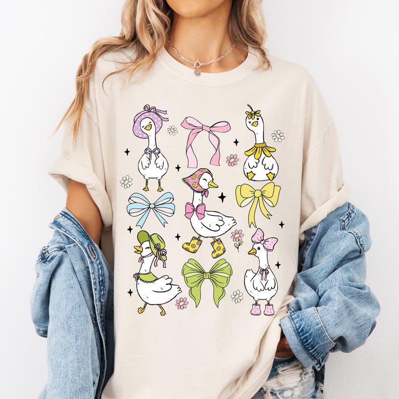 Puede incluir: Una camiseta de color crema con un gr&aacute;fico de seis gansos de dibujos animados que llevan sombreros y lazos. Los gansos son blancos con picos y patas amarillas. Los sombreros son de varios colores y estilos, incluyendo un sombrero de flor rosa, un sombrero verde con un lazo y un sombrero azul con un lazo. Los lazos tambi&eacute;n son de varios colores y estilos, incluyendo un lazo rosa, un lazo azul y un lazo amarillo. Los gansos est&aacute;n rodeados de peque&ntilde;as flores y estrellas.