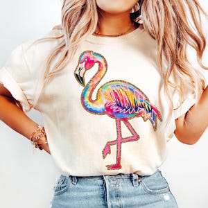 May include: Cream-colored t-shirt featuring a vibrant, watercolor-style flamingo. The flamingo has a rainbow of colors, outlined in gold glitter. The bird is standing on one leg, with a pink leg and beak.