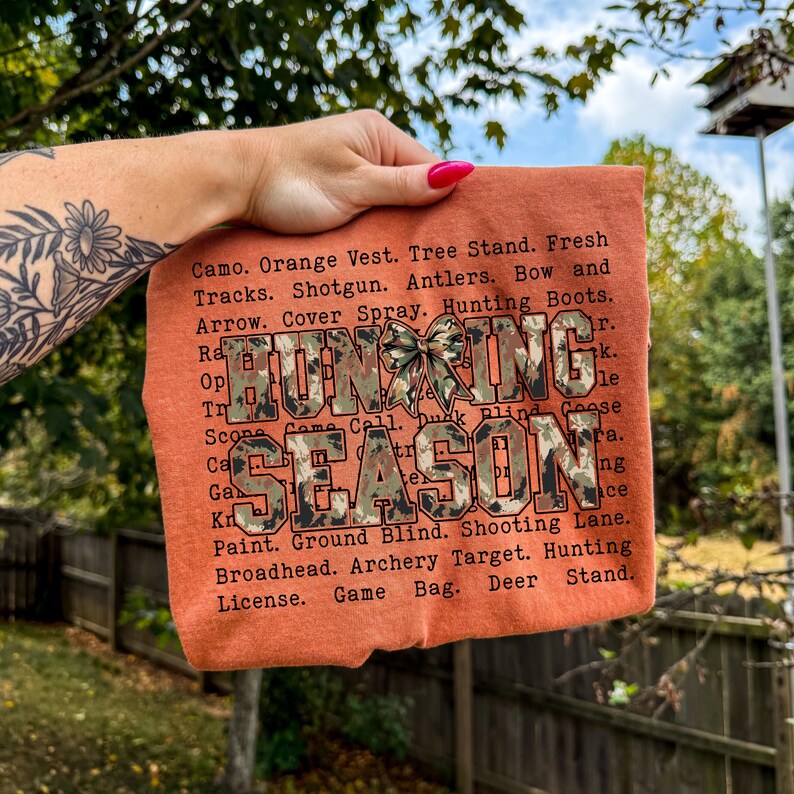 May include: An orange t-shirt with the words "HUNTING SEASON" in a camouflage design. The shirt lists hunting-related terms such as "Shotgun," "Antlers," and "Game Bag."
