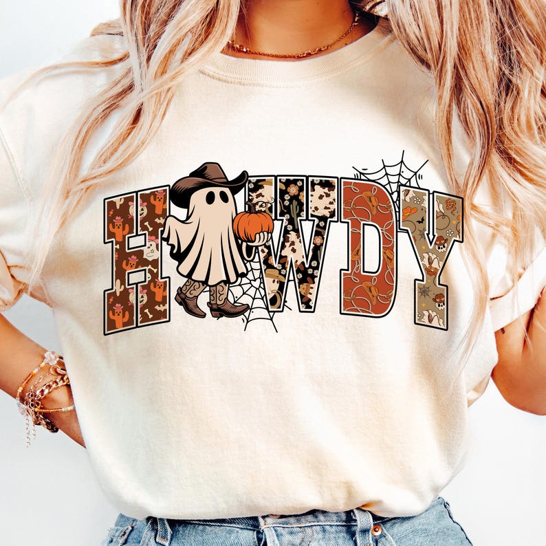 Puede incluir: Camiseta color crema con la palabra "HOWDY" en un dise&ntilde;o de tem&aacute;tica occidental. La "H" y la "Y" est&aacute;n decoradas con patrones florales y de cactus, mientras que la "O" y la "W" tienen un estampado de piel de vaca. Un fantasma con sombrero y botas de vaquero sostiene una calabaza.