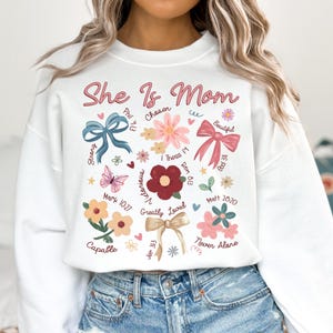 K&ouml;nnte beinhalten: Wei&szlig;er Pullover mit dem Text "She Is Mom" in rosa Schrift, umgeben von einem Blumendesign mit Schleifen, Schmetterlingen und Bibelversen.