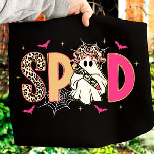 Pu&ograve; includere: T-shirt nero con la scritta "SPOD" in stampa leopardata, beige e rosa. Un fantasma con un fiocco leopardato e una matita al centro. Pipistrelli e stelle rosa circondano il testo e il fantasma.