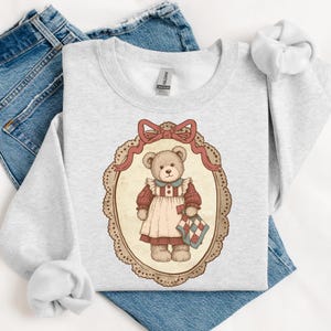 Op de afbeelding: Een lichtgrijs sweatshirt met een vintage illustratie van een teddybeer in een ovale lijst, compleet met een rode strik. De beer draagt een jurk en houdt een quilt vast. Het sweatshirt ligt op een blauwe jeans.