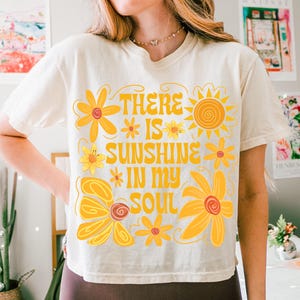 May include: A white t-shirt with a yellow floral design and the text "There is sunshine in my soul".