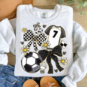 May include: A light gray sweatshirt featuring a soccer-themed design. The design includes a soccer ball, a jersey with the number 7, bows, daisies, and a soccer glove. The design incorporates black, white, and gold accents.