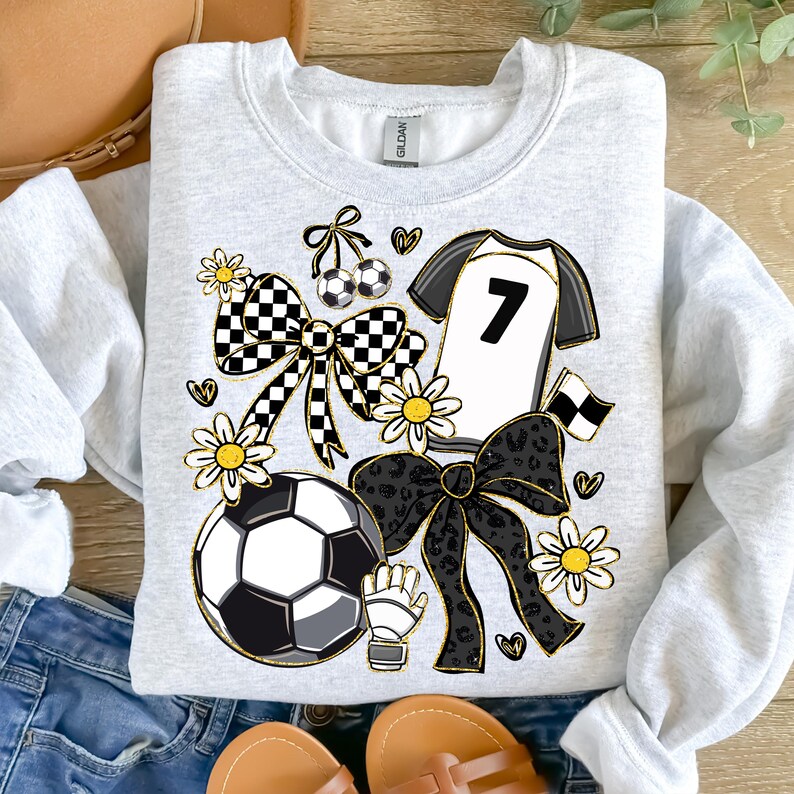 May include: A light gray sweatshirt featuring a soccer-themed design. The design includes a soccer ball, a jersey with the number 7, bows, daisies, and a soccer glove. The design incorporates black, white, and gold accents.