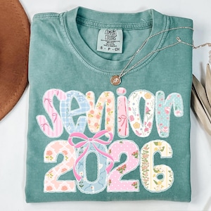 Puede incluir: Una camiseta Comfort Colors verde azulado con la palabra "Senior 2026" en un dise&ntilde;o de patchwork. El patchwork incluye patrones florales y de lazo. Un collar dorado con un colgante est&aacute; en la camisa.