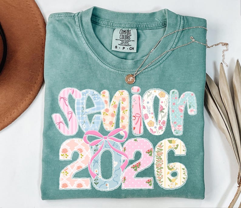 Puede incluir: Una camiseta Comfort Colors verde azulado con la palabra "Senior 2026" en un dise&ntilde;o de patchwork. El patchwork incluye patrones florales y de lazo. Un collar dorado con un colgante est&aacute; en la camisa.