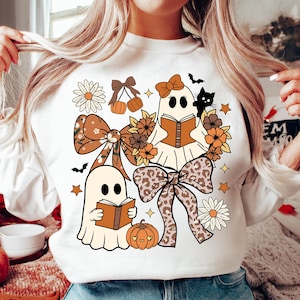Op de afbeelding: Witte sweatshirt met een Halloween-thema. Het ontwerp toont twee spookfiguren die boeken lezen, omringd door strikken, pompoenen, bloemen, sterren en een zwarte kat. De afbeelding is in tinten bruin, oranje en cr&egrave;me.