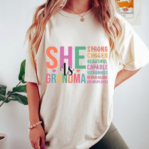 May include: A white t-shirt with the text "SHE is GRANDMA" in colorful letters. The text is surrounded by other words in smaller letters, including "strong", "chosen", "beautiful", "capable", "victorious", "never alone", and "greatly loved".
