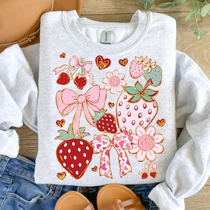 Op de afbeelding: Een grijs sweatshirt met een kleurrijk ontwerp van aardbeien, strikken en harten. Het ontwerp is in retro-stijl met een roze, rode en groene kleurstelling.