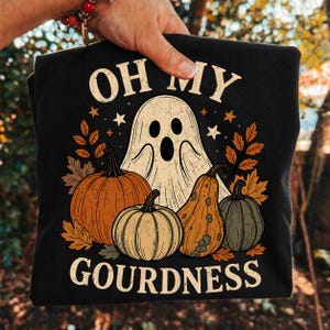 May include: A black t-shirt featuring a graphic print of a white ghost, orange and green pumpkins, and autumn leaves. The text "OH MY GOURDNESS" is printed in a cream-colored font above and below the central illustration of the ghost and gourds.