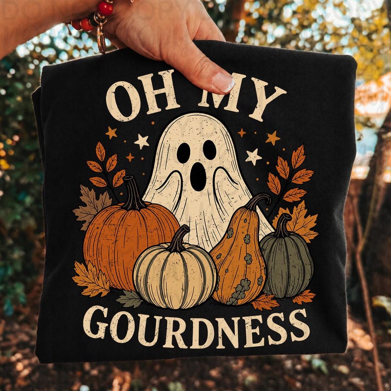 May include: A black t-shirt featuring a graphic print of a white ghost, orange and green pumpkins, and autumn leaves. The text "OH MY GOURDNESS" is printed in a cream-colored font above and below the central illustration of the ghost and gourds.