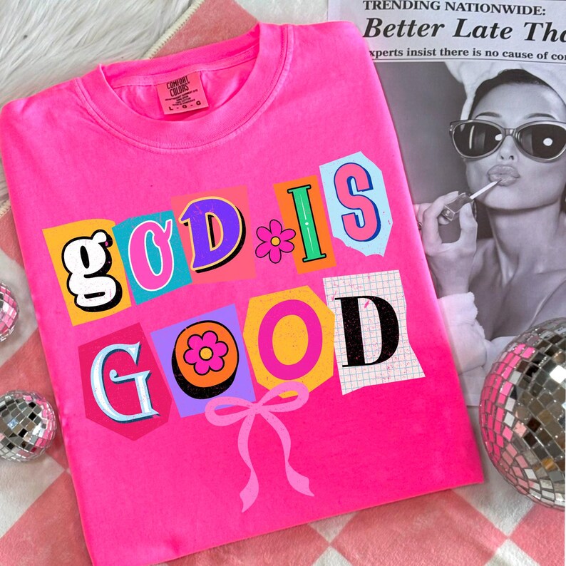 May include: A bright pink Comfort Colors sweatshirt with the phrase "GOD IS GOOD" in a colorful, collage-style design. The design includes a flower and a pink bow. A black and white image of a woman applying lipstick is in the background.
