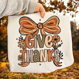 May include: A cream-colored decorative pillow with the words "GIVE THANKS" in a fall-themed design. The design includes a large bow, flowers, and leaves in shades of orange, brown, and beige.