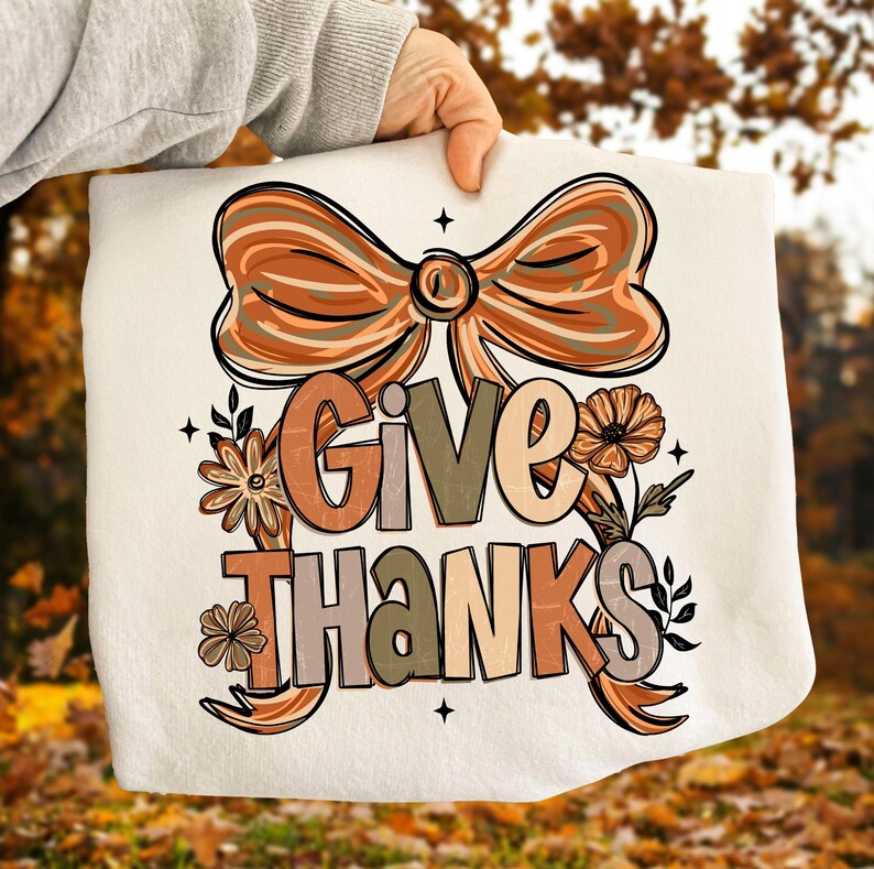 May include: A cream-colored decorative pillow with the words "GIVE THANKS" in a fall-themed design. The design includes a large bow, flowers, and leaves in shades of orange, brown, and beige.