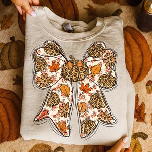 May include: A beige sweatshirt featuring a large bow design with a pumpkin and fall leaf pattern. The bow has a leopard print center and is decorated with orange and brown leaves and flowers. The sweatshirt is folded and held by hands.