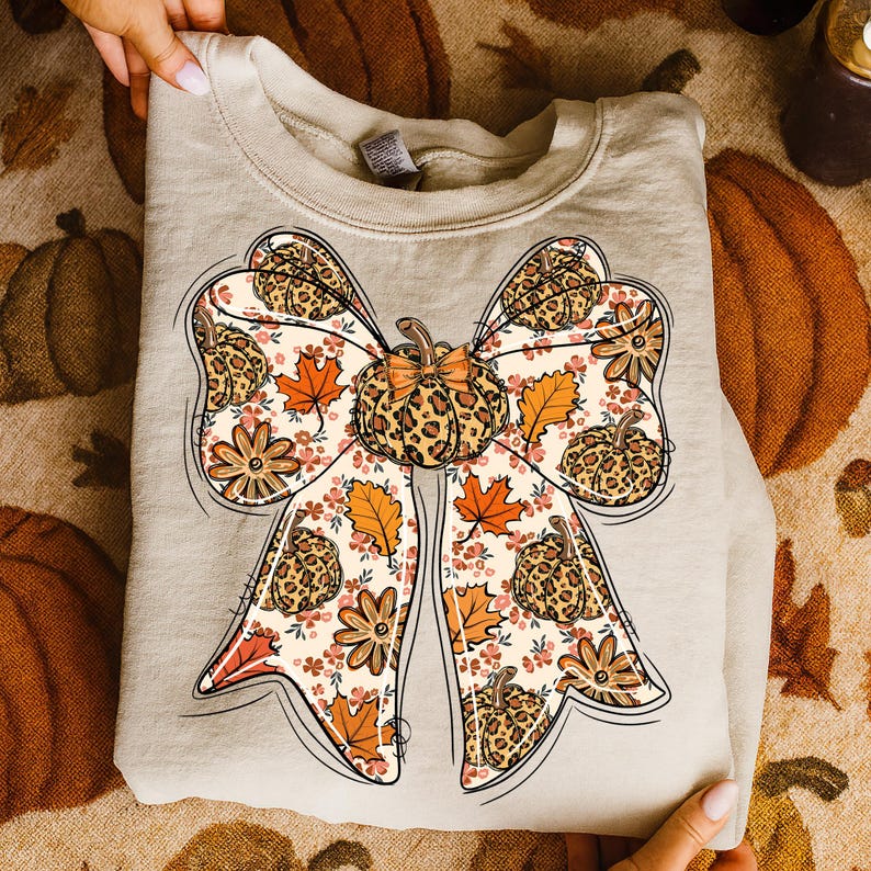 May include: A beige sweatshirt featuring a large bow design with a pumpkin and fall leaf pattern. The bow has a leopard print center and is decorated with orange and brown leaves and flowers. The sweatshirt is folded and held by hands.