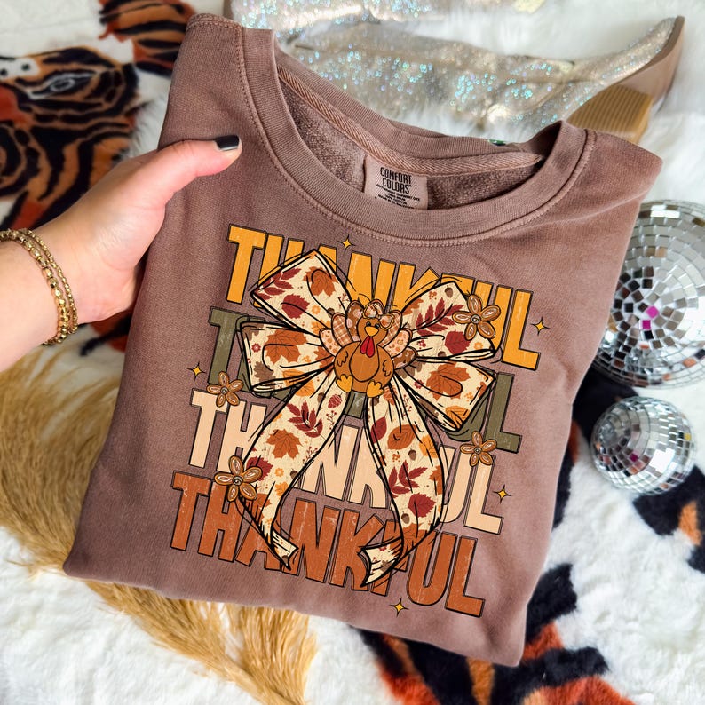 May include: A brown sweatshirt with the word "THANKFUL" in orange and yellow block letters. A large bow with a turkey in the center is the focal point. The bow has a fall leaf pattern. The sweatshirt is a Comfort Colors brand.